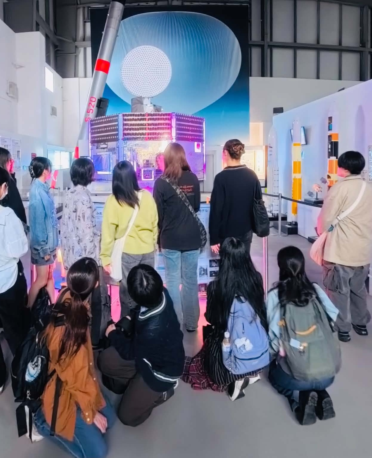 JAXA相模原キャンパス・宇宙科学探査交流棟の展示室。S-520観測ロケットと探査機のモデルを、十数名の学生が取り囲んで見ている